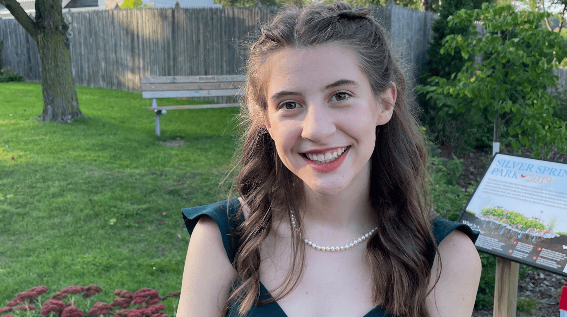 Young woman smiling outdoors, wearing a dark green dress and a pearl necklace, with long wavy brown hair partially braided. She stands in front of a park sign, a bench, and greenery on a sunny day.